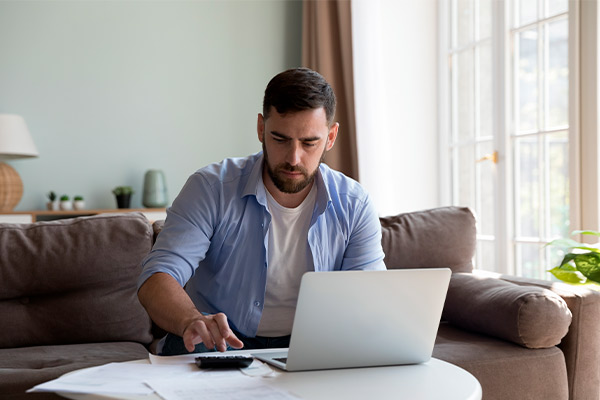 A man on a couch uses a laptop and reviews papers about first-time home buyer programs available in South Texas.