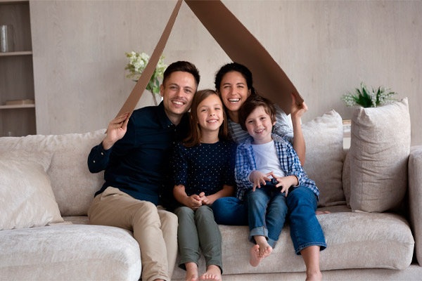 Family portrait smiling parents with kids sitting on couch under cardboard roof, happy mother and father with little daughter and son playing funny game in new house, mortgage and relocation concept